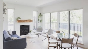 A modern living room with a fireplace, a dining area with a glass table, and large windows offering a view of the outdoors.