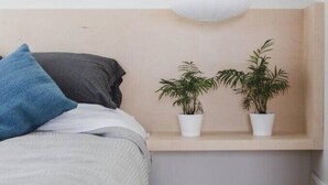 A bedroom with a bed, a bedside table with two potted plants, a white pendant light, and a wooden headboard.