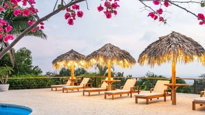 Een zwembadterras met ligbedden en rieten parasols, omringd door weelderige begroeiing en een helder uitzicht op de oceaan.