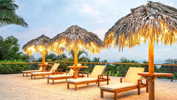 Een terras aan het strand met houten ligbedden en rieten parasols, met uitzicht op een mooi landschap.