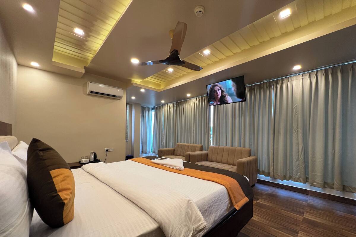 A modern hotel room with a large bed, a seating area with two sofas, a flat-screen TV mounted on the wall, and a ceiling fan.