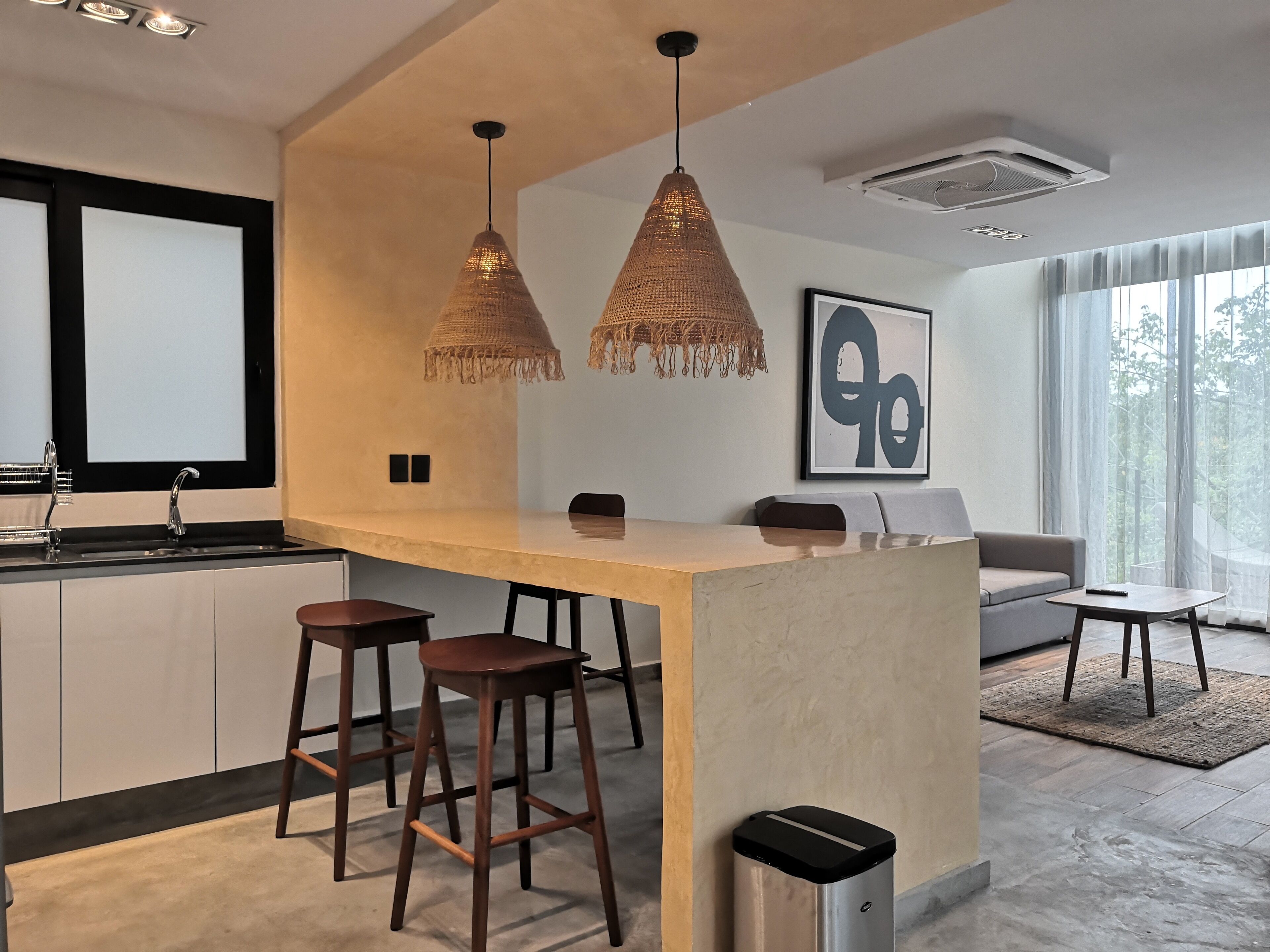A modern kitchen with a central island, bar stools, pendant lights, a large window, and a sofa in the living area.