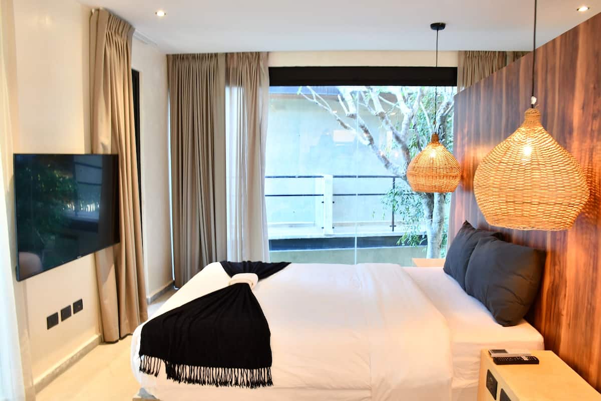 A modern hotel room with a large bed, a flat-screen TV, and a balcony with a view of trees.