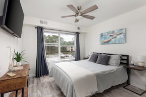 Quarto com cama, ventoinha no teto, televisão, janela com cortinas, mesa de cabeceira com abajur e um quadro na parede.