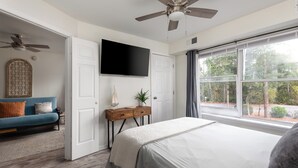 A bedroom with a bed, a TV mounted on the wall, a ceiling fan, a window with curtains, a wooden nightstand, and a blue sofa.