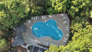 An aerial view of a swimming pool surrounded by lounge chairs and a covered patio area, with trees in the background.