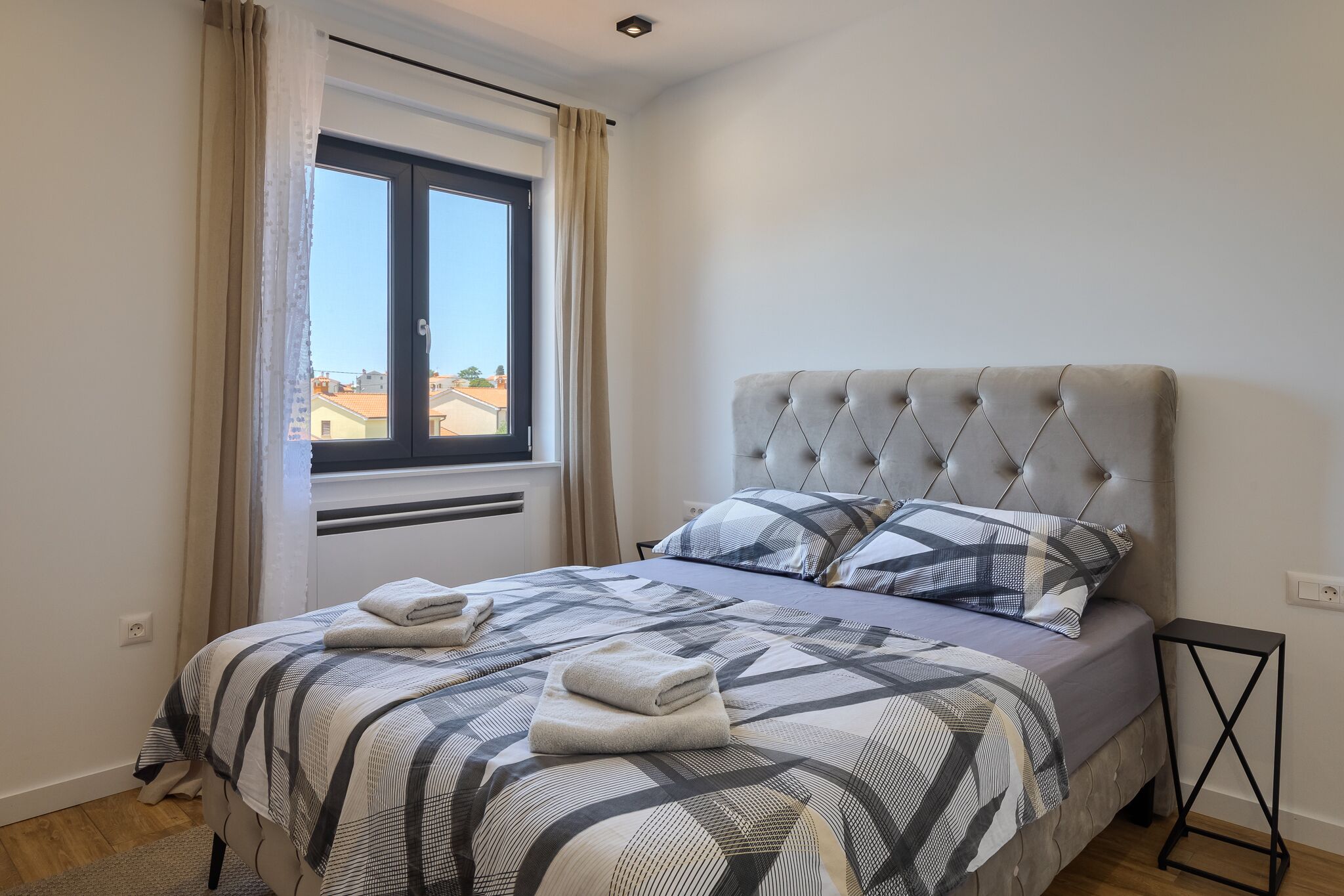 A neatly made bed with a checkered bedspread and two pillows, a window with curtains, and a small black side table.