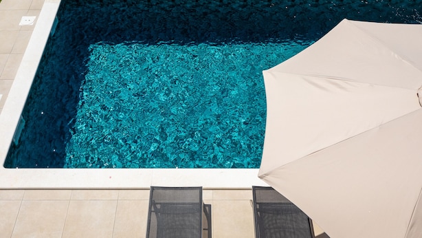 Ein Swimmingpool mit klarem, blauem Wasser, umgeben von Fliesen und zwei Liegestühlen mit einem beigen Sonnenschirm.
