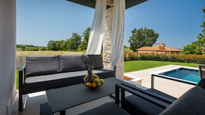 Eine Terrasse mit einer schwarzen Couch, einem Tisch mit Obstschale und Blick auf Schwimmbad und Garten.