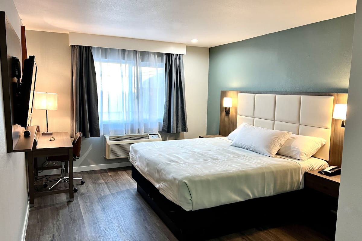 Quarto de hotel com cama, escrivaninha, cadeira, luminária e aparelho de ar condicionado.
