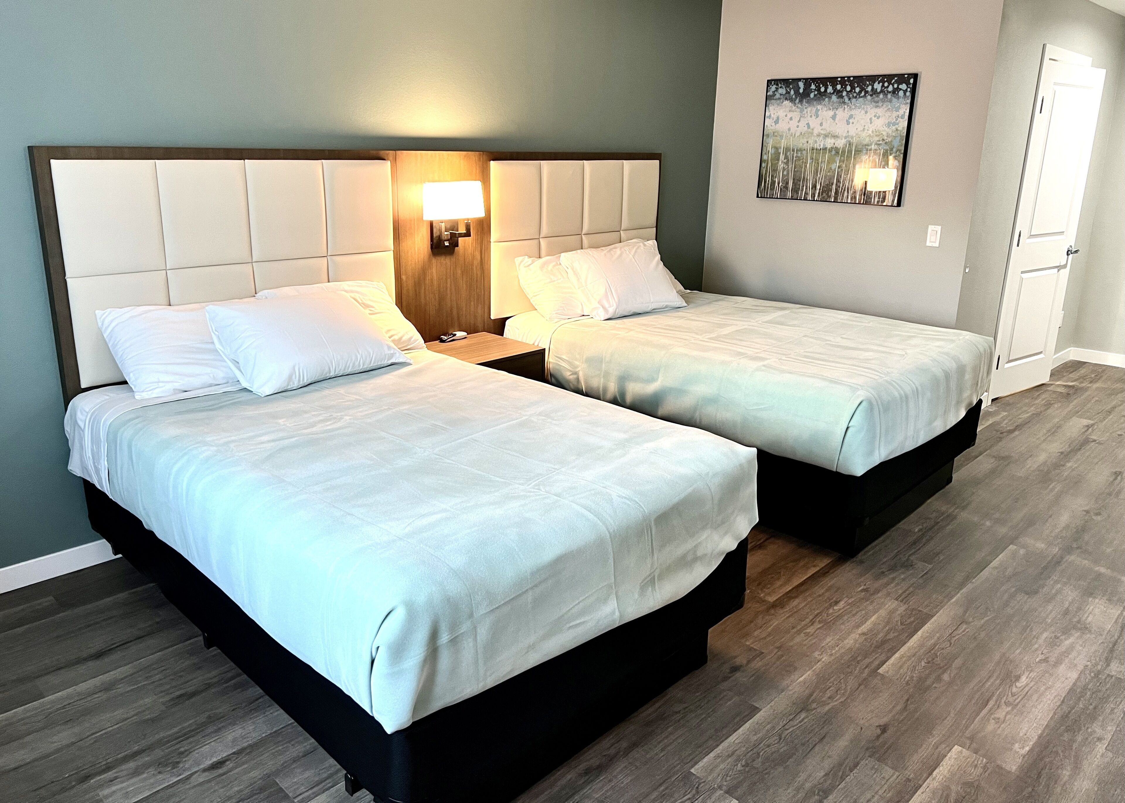 A hotel room with two beds, a nightstand with a lamp, a painting on the wall, and a wooden floor.
