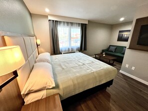A hotel room with a bed, a nightstand with a lamp, a small table, a sofa, and a window with curtains.