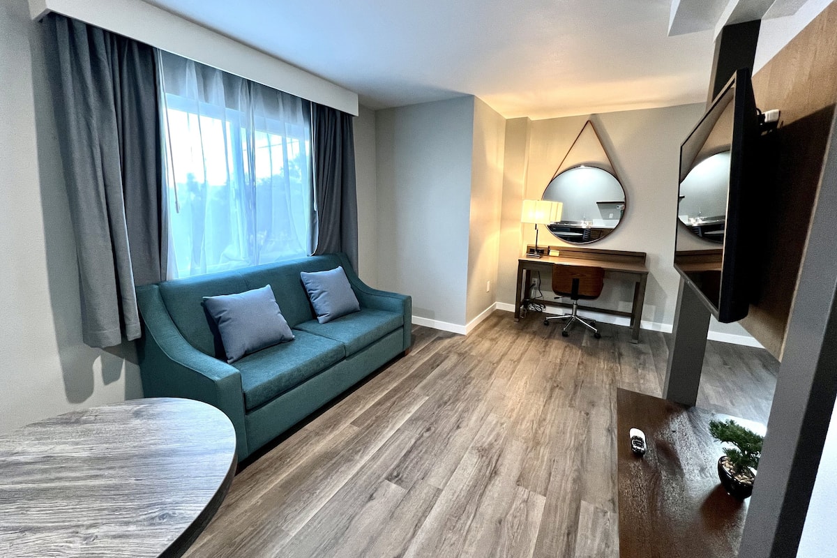Quarto de hotel moderno com sofá verde-água, piso de madeira, uma mesa redonda e uma escrivaninha com abajur.