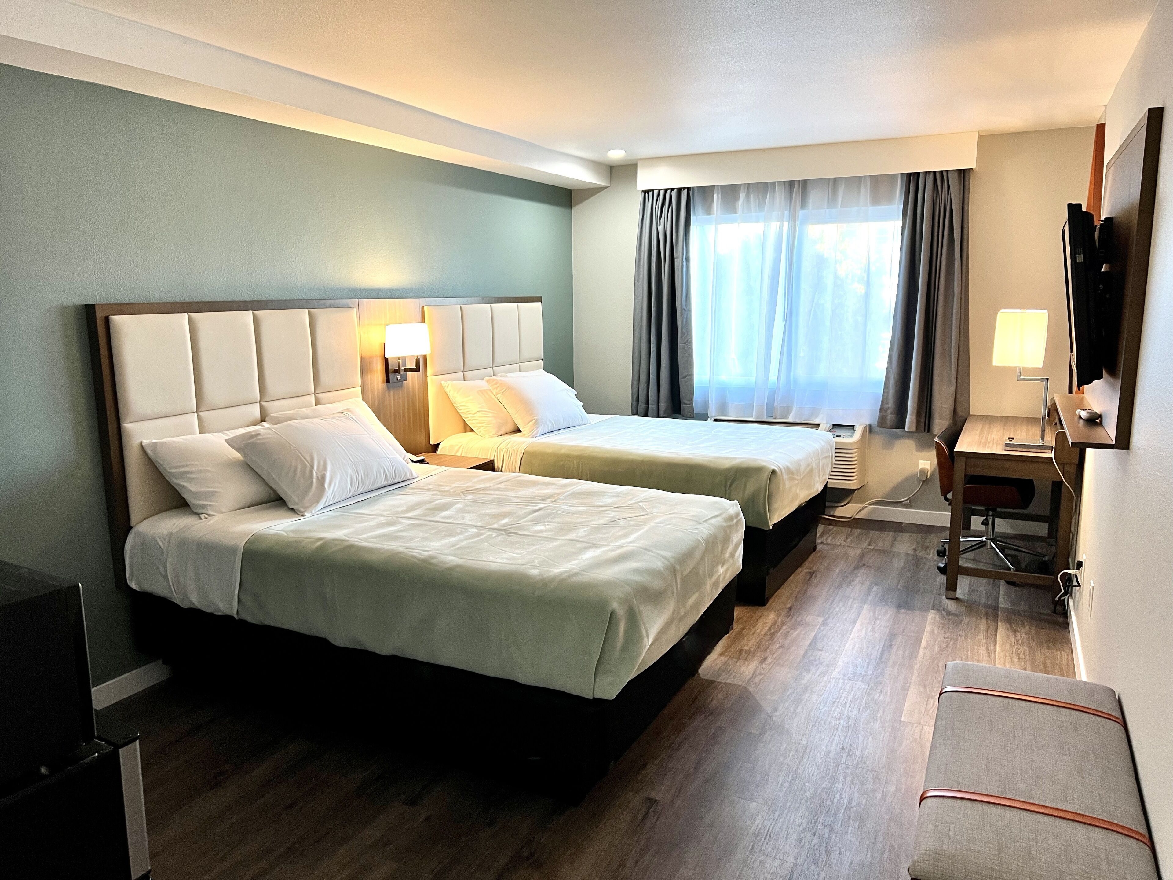 Hotellihuone, jossa on kaksi sänkyä, työpöytä, tuoli, televisio ja ikkuna, jossa on verhot.