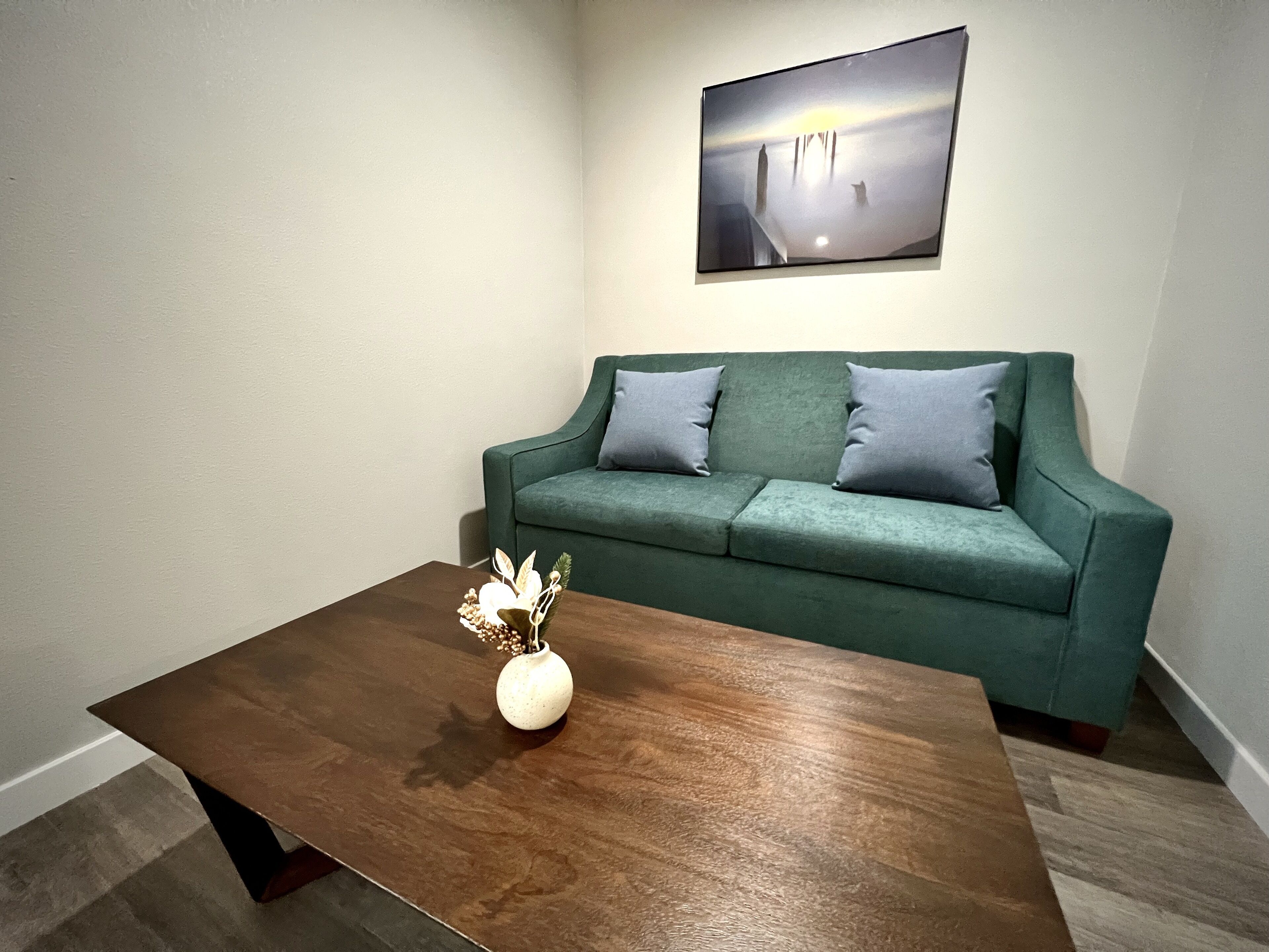 A living room with a green sofa, two cushions, a wooden coffee table with a vase of flowers, and a framed picture on the wall.