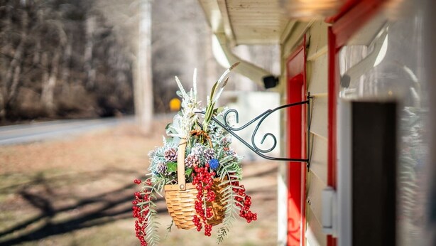A wicker basket with greenery and red berries hanging from a metal hook on a building's exterior. - Quaint Cabin By The Creek Privat (Pipestem)