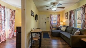A cozy living room with a sofa, a dining table with chairs, a ceiling fan, and patterned curtains. - Quaint Cabin By The Creek Privat (Pipestem)