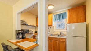 A compact kitchen with wooden cabinets, a white refrigerator, a coffee maker, and a dining area with a table and chairs. - Quaint Cabin By The Creek Privat (Pipestem)