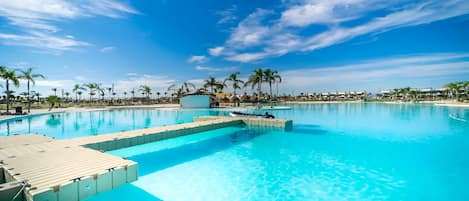 A large outdoor swimming pool with clear blue water, surrounded by palm trees and a clear sky.