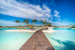 A wooden boardwalk leading to a pool with clear blue water, surrounded by palm trees and a white sandy area.