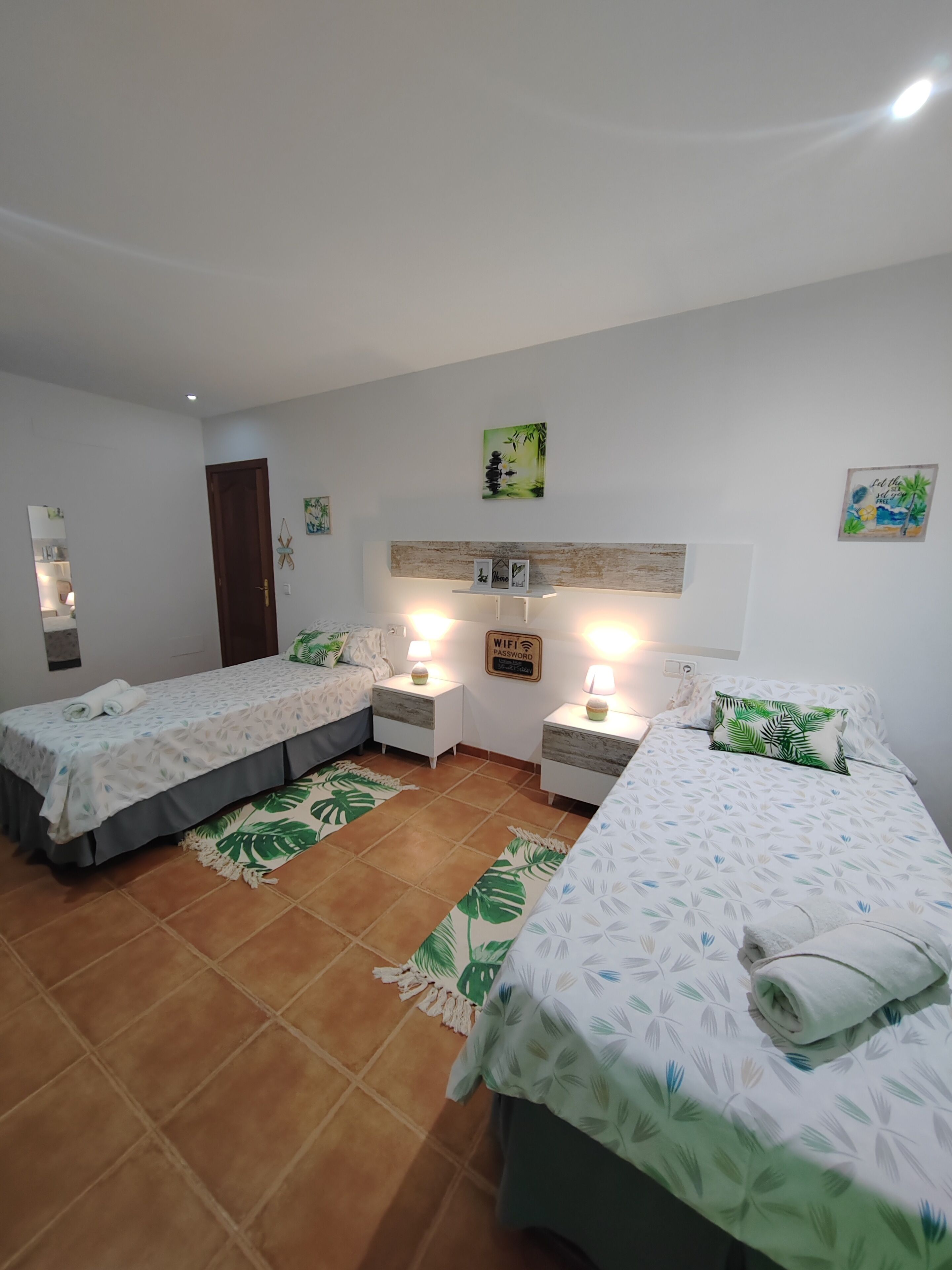 A room with two beds, each with a nightstand and a lamp. There are wall-mounted shelves with decorative items and a sign indicating WiFi availability.