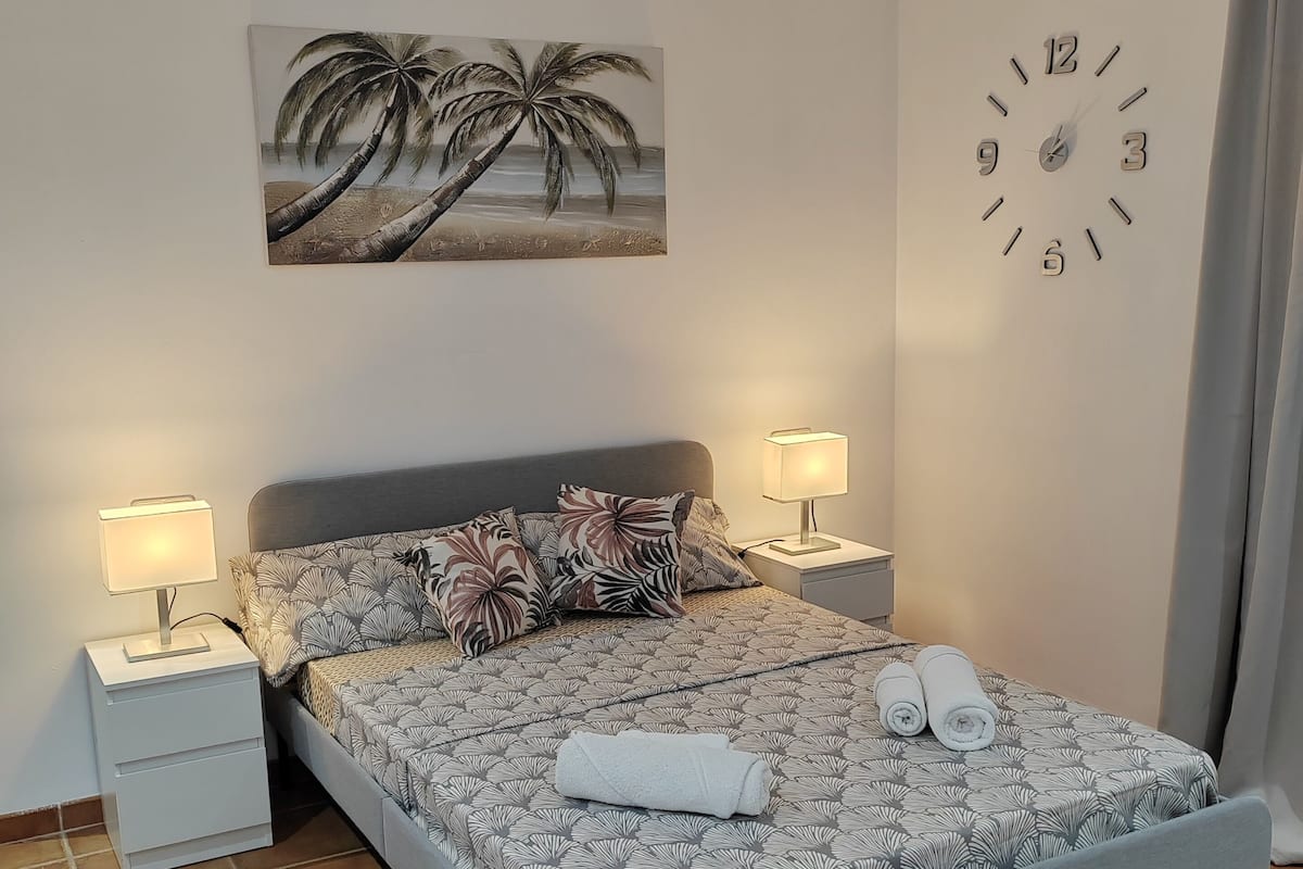 A neatly arranged bedroom with a bed, two bedside tables, a wall clock, and a tropical-themed painting.