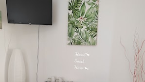 Ein kompakter Wohnraum mit Wand-Fernseher, kleinem Schreibtisch mit Stuhl, kompaktem Kühlschrank und Topfpflanze.