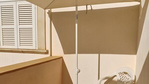 Ein kleiner Außenbereich mit einer Terrasse, einem weißen Tisch und Stühlen, einem weißen Sonnenschirm und einem Fenster mit weißen Rollläden.