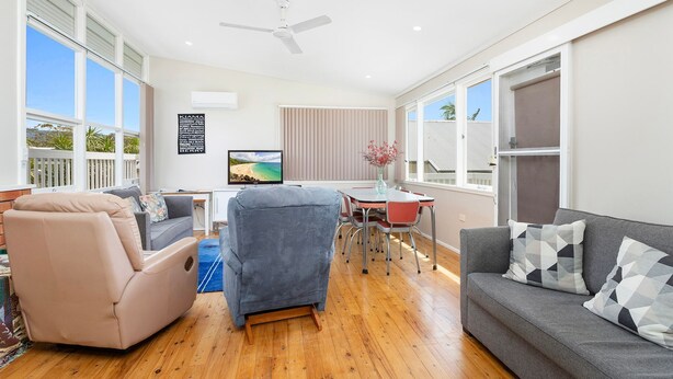 A spacious living room with a large window, a ceiling fan, a flat-screen TV, and a dining area with a table and chairs. - Jack's Place - Cozy Retreat (Gerringong)