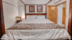 A bedroom with a large bed, wooden headboard, two framed pictures on the wall, a nightstand with a vase of flowers, and a wooden door.