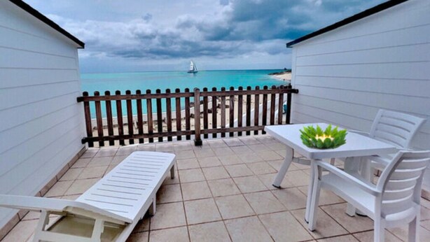 Un balcon avec vue sur la plage et l’océan, une table blanche ornée d’une plante et deux chaises longues blanches.