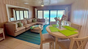 Un salon avec un canapé, une table à manger et des chaises, une grande fenêtre donnant sur l’océan et un ventilateur de plafond.
