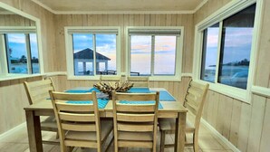 A dining area with a wooden table and chairs, large windows with views of the sea and a building, and a decorative centerpiece. - Sea La Vie - Schooner's Landing (Man-O-War Cay)