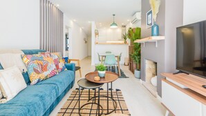 Uma sala de estar moderna com um sofá azul, uma mesa de centro e uma TV. Há uma área de jantar com mesa e cadeiras, e uma cozinha ao fundo.