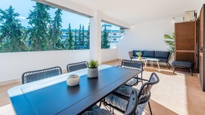 Uma área de refeições externa moderna com uma mesa e cadeiras pretas, um vaso com planta e vista para árvores e edifícios.