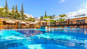 Piscina com espreguiçadeiras e guarda-sóis, rodeada por edifícios e árvores.