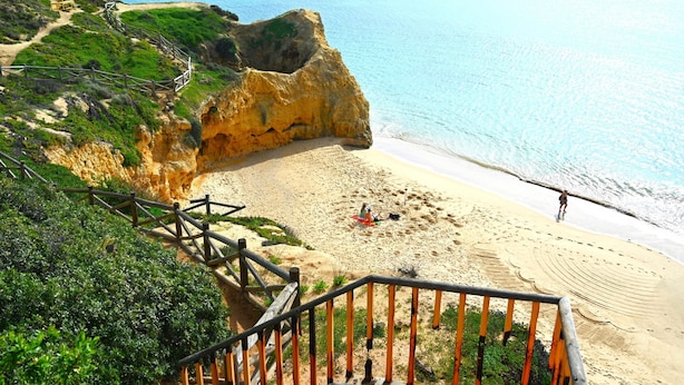 Uma praia costeira com uma escadaria de madeira que desce até a orla, areal arenoso e vista clara do oceano.