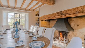 Comedor con una larga mesa de madera dispuesta para una comida, sillas blancas y una urna grande, azul y blanca. Se ve una chimenea de piedra con fuego encendido al fondo.