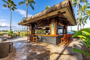 A tiki bar with a thatched roof, wooden chairs, and a stone counter.