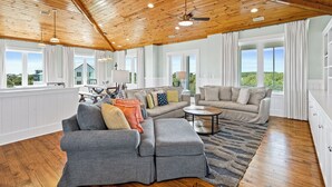 A spacious living room with a high ceiling, wooden beams, and large windows offering a view of the outdoors.