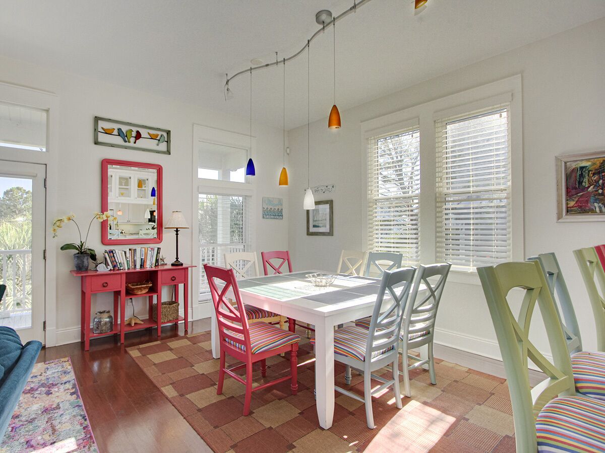 A dining area with a white table and chairs, a pink console table with books and a lamp, and a window with blinds.