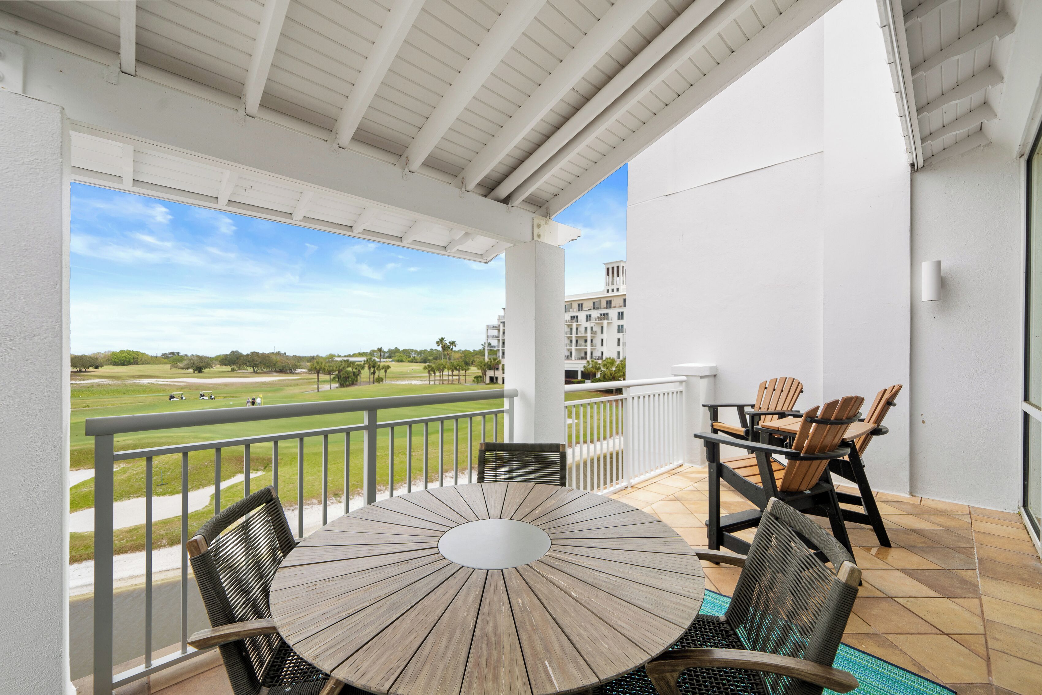 A balcony with a round table and chairs, a view of a golf course, and a white building in the background.