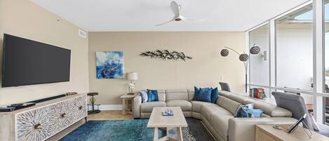 A modern living room with a flat-screen TV, a sectional sofa, a coffee table, and a large window.