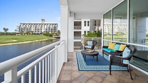 Un balcon avec vue sur une rivière et un bâtiment situé de l’autre côté. On y trouve un canapé en osier avec des coussins et une petite table sur laquelle est posé un saladier.
