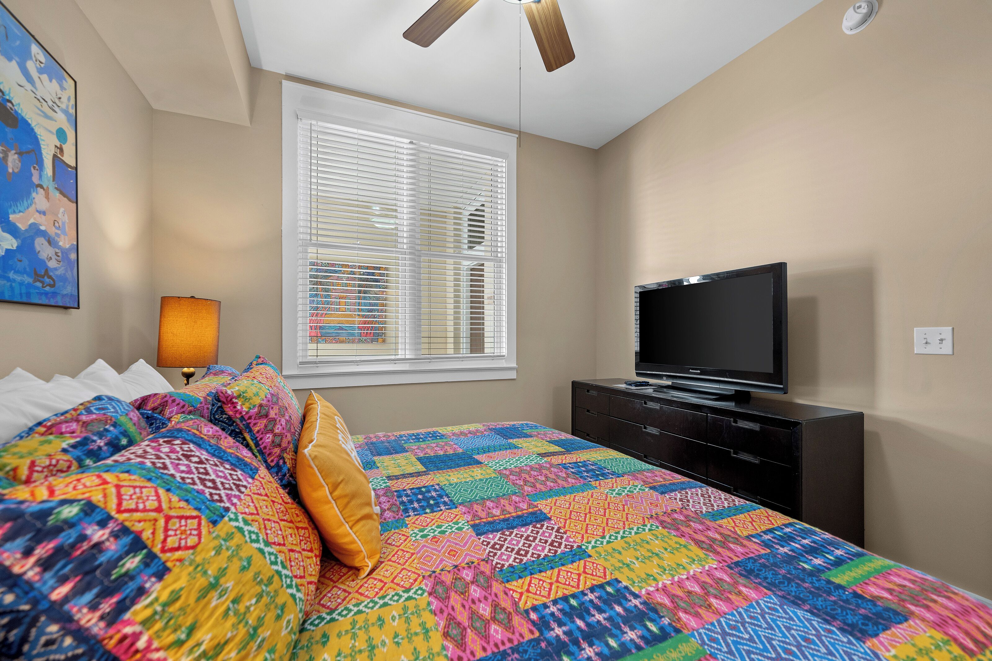 A bedroom with a bed, a TV, a window with blinds, a ceiling fan, and a painting on the wall.