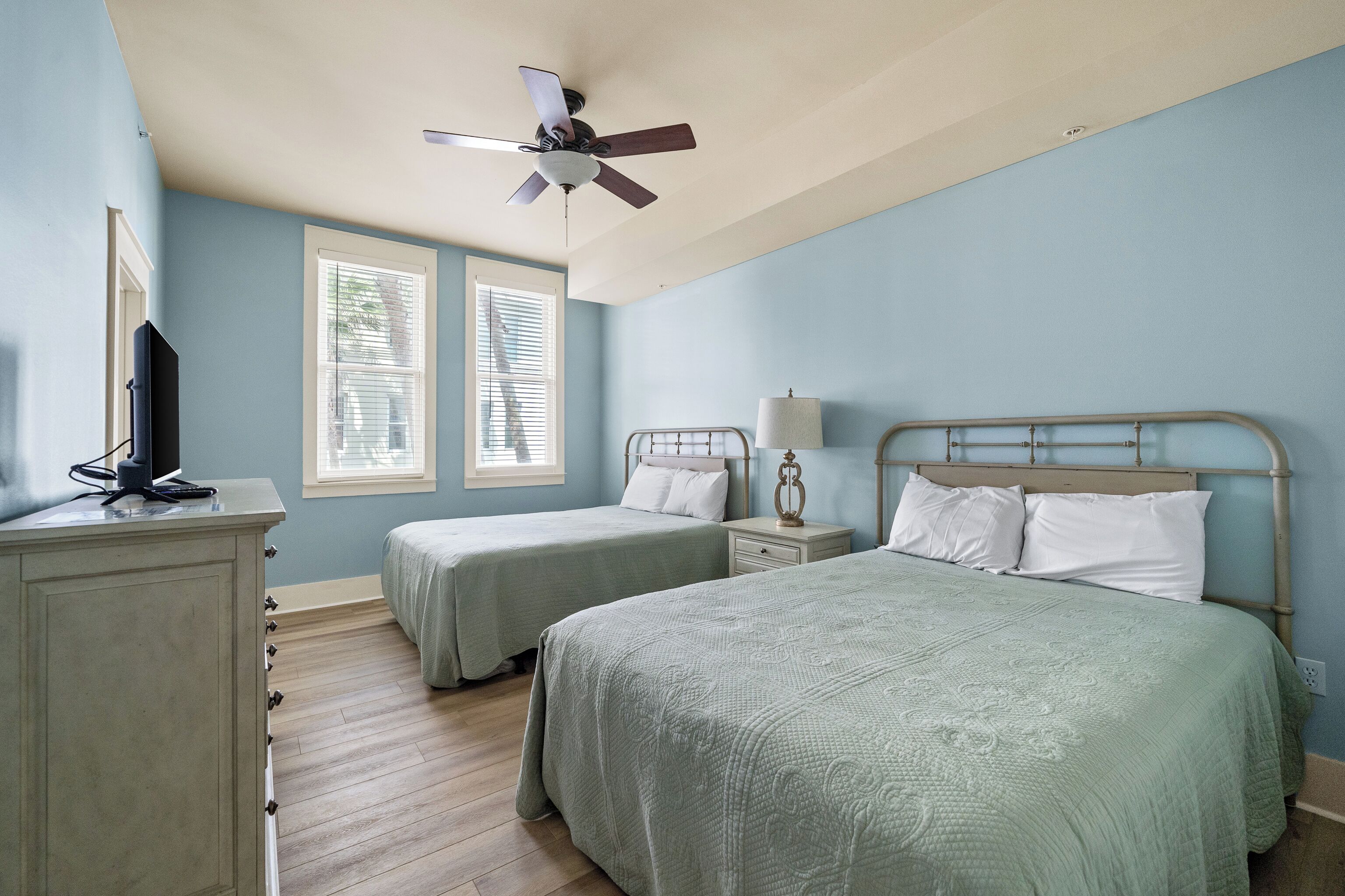 Une chambre avec deux lits, un ventilateur de plafond, une table de chevet avec une lampe et une commode.