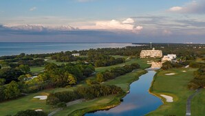 An aerial view of a golf course with a large building, likely a hotel, in the background.