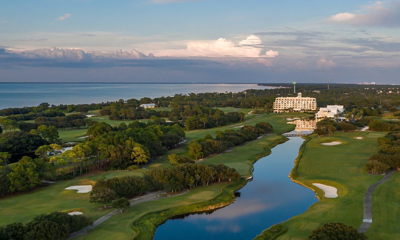 An aerial view of a golf course with a large building, likely a hotel, in the background.