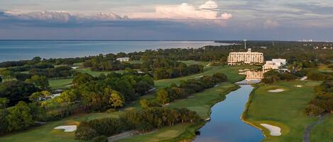 An aerial view of a golf course with a large building, likely a hotel, in the background.