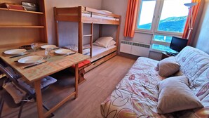A dormitory room with bunk beds, a desk with plates and cutlery, a bed with a floral duvet, and a window with curtains.